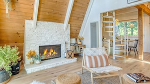 A cozy living room with a fireplace, wooden beams, and a staircase leading to an upper level. - A-frame Retreat by Avantstay Modern Interiors 2 Decks Views (Skyforest)