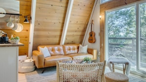 A cozy loft-style room with a wooden ceiling, a leather sofa, a guitar, and a large window offering a view of the outdoors. - A-frame Retreat by Avantstay Modern Interiors 2 Decks Views (Skyforest)