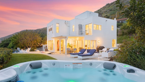 A modern two-story house with a hot tub in the foreground, a patio with lounge chairs, and a view of a hillside. - Stardust by Avantstay Views Hot Tub Pool Table (Malibu)