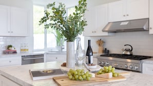 A kitchen with white cabinets, a marble countertop, a vase with green leaves, a bottle of wine, and a cutting board with grapes. - Stardust by Avantstay Views Hot Tub Pool Table (Malibu)