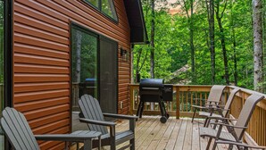 A deck with Adirondack chairs and a grill, surrounded by a wooden deck and trees.