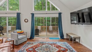 A spacious living room with a large window, a flat-screen TV, a grey sofa, a patterned rug, and a wooden bench.