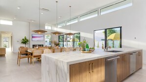 A modern kitchen with a large island, wooden cabinets, and stainless steel appliances. There is a dining area with a wooden table and chairs, and a large window providing natural light.