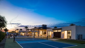 A tennis court with a net, surrounded by modern buildings with large windows and outdoor seating areas.