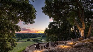 A circular outdoor seating area with chairs and a table, surrounded by rocks and trees, overlooking a vineyard and distant hills.