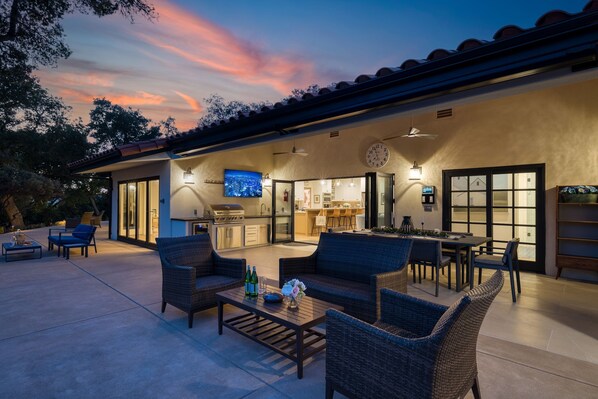 An outdoor patio area with wicker furniture, a dining table, and a kitchen visible through glass doors. - Rancho Robles by Avantstay Vineyard Villa w Views Pool Privacy (Paso Robles)