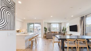 Uma cozinha moderna com uma ilha central, uma sala de jantar com mesa escura e cadeiras de madeira, e uma sala de estar com sofá e televisão.