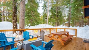 A deck with blue Adirondack chairs, a fire pit, and a wooden picnic table surrounded by snow and trees.