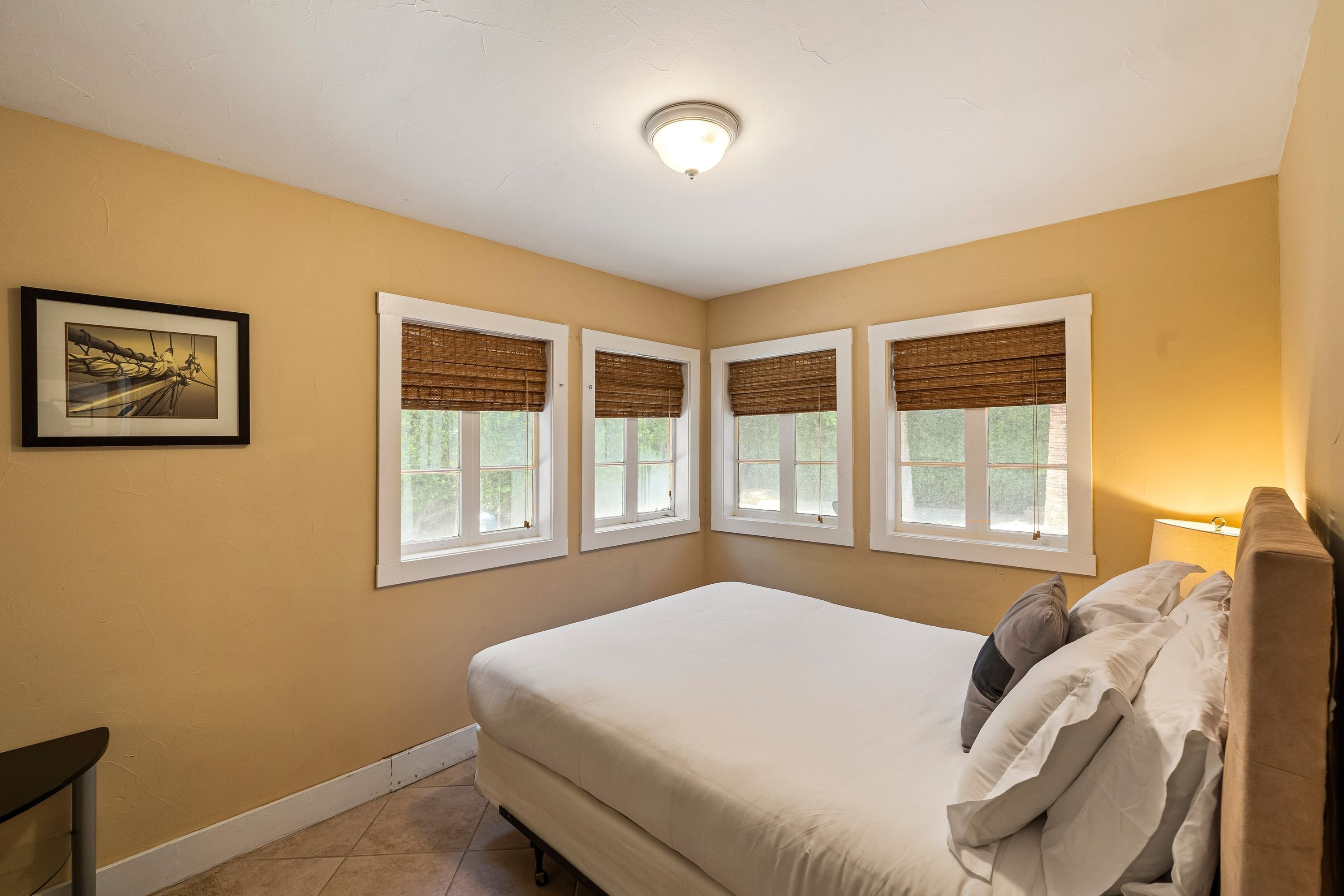 A bedroom with a bed, four windows with blinds, a ceiling light, and a framed picture on the wall.