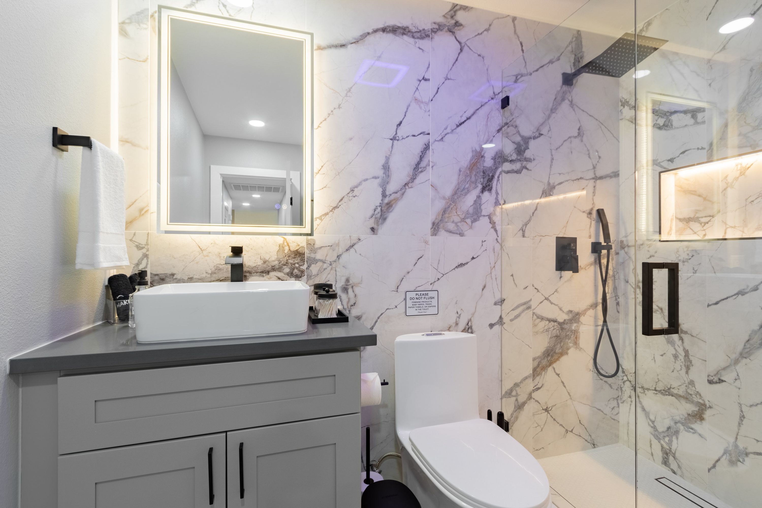 A modern bathroom with a marble backsplash, a large mirror, a white sink, and a glass-enclosed shower.
