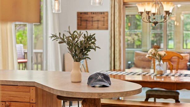 Una cocina con encimera de madera, un florero con plantas verdes y una gorra. Hay una araña sobre la encimera y un letrero en la pared que dice 'WINE TASTING'.