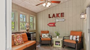 Una habitación acogedora con techo de madera, un sofá con cojines estampados, dos sillones de mimbre con cojines anaranjados, una mesita con una lámpara y un cartel en la pared que indica 'LAKE' y 'Cabins'.