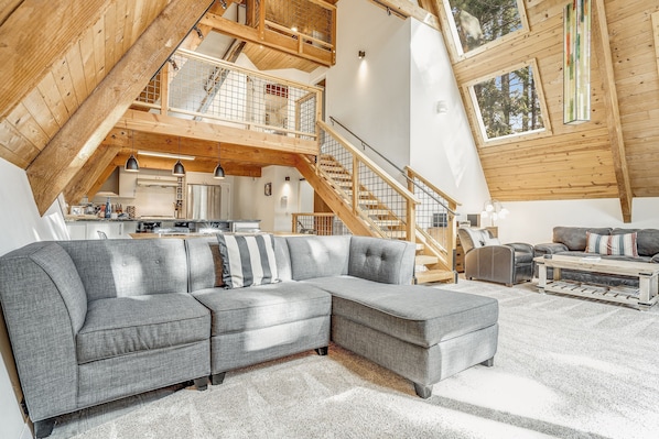 Un loft amplio con un gran sofá seccional gris, vigas de madera y un entrepiso con una escalera.