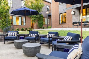 An outdoor seating area with multiple chairs and round tables, a large umbrella, and a brick building in the background. - The Magnolia 10 - Avantstay Germantown w Private Balcony (Nashville)