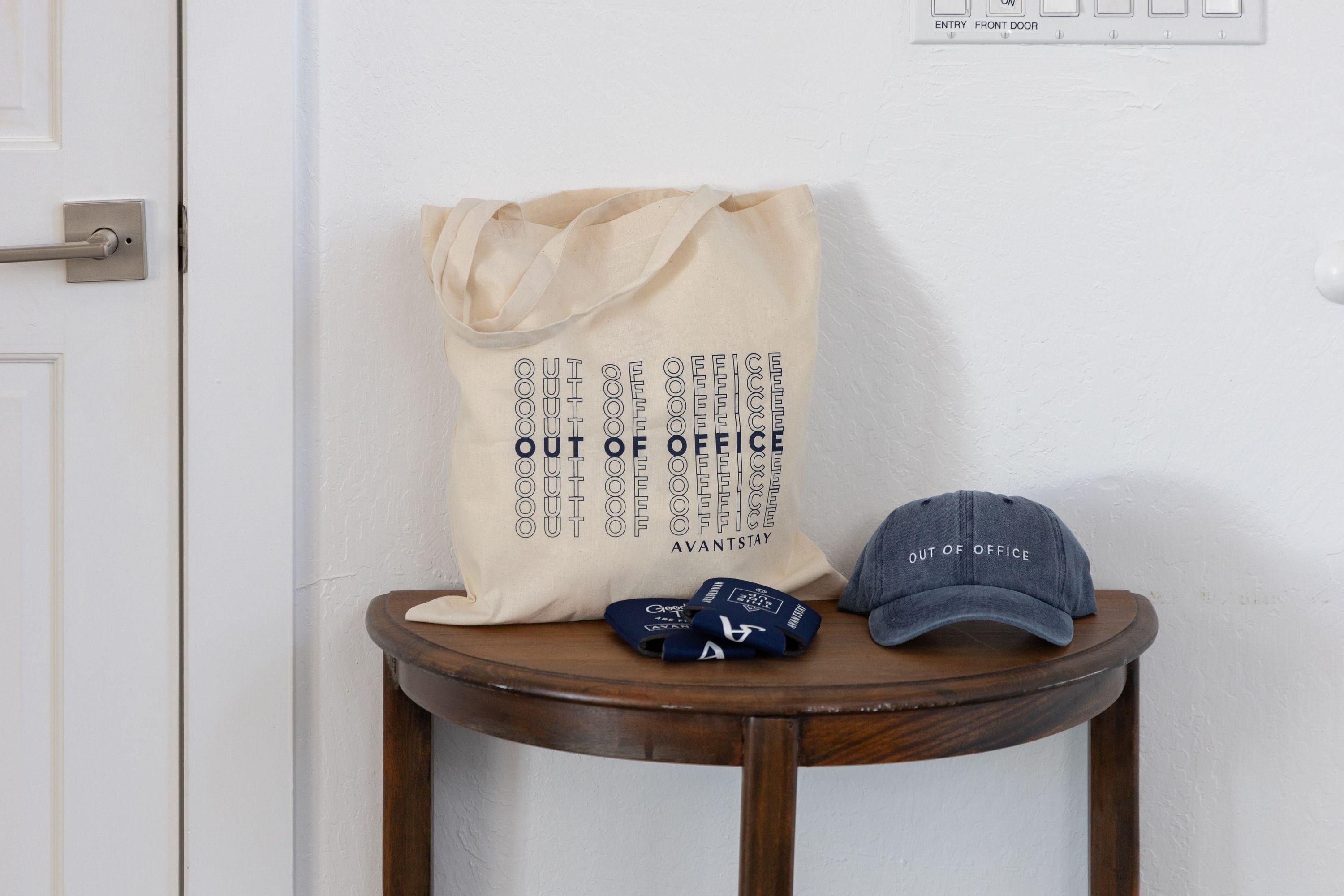 A beige tote bag with "OUT OF OFFICE" printed on it, placed on a wooden table next to a blue cap and a pair of blue slippers.