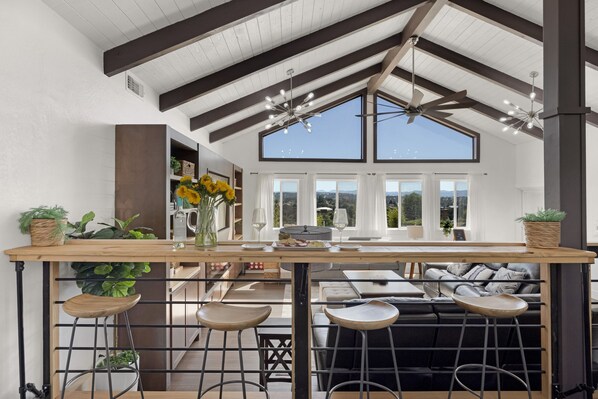 Una cocina amplia con techo alto, vigas de madera y ventanas grandes. Hay taburetes y una barra con plantas.