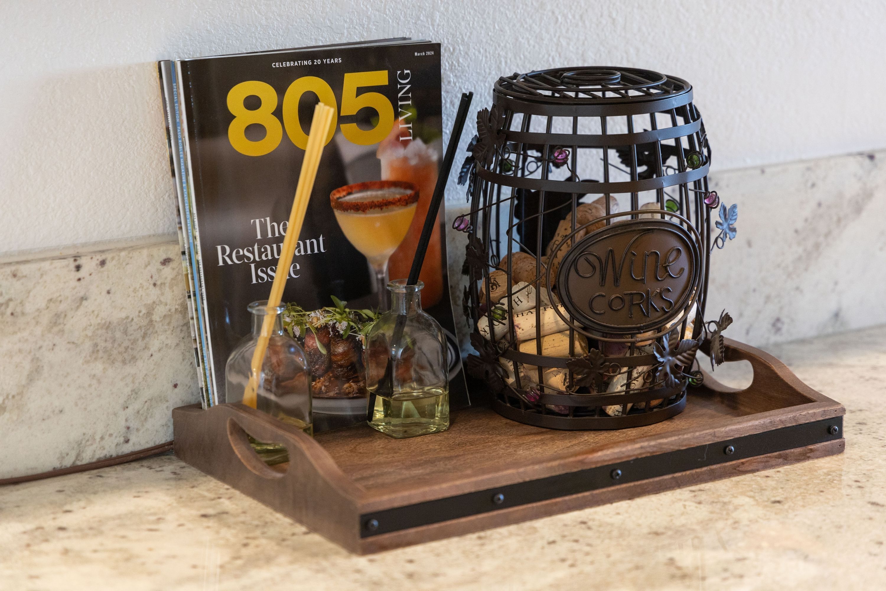 A magazine titled "805 Living The Restaurant Issue" on a wooden tray with a wine barrel and a glass bottle.