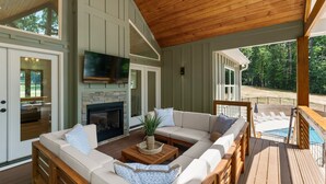 A covered outdoor living area with a wooden deck, a fireplace, a large sofa, and a television.