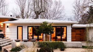 A modern house with a snow-covered roof, a well-lit entrance, and a garden with various plants and a small tree.