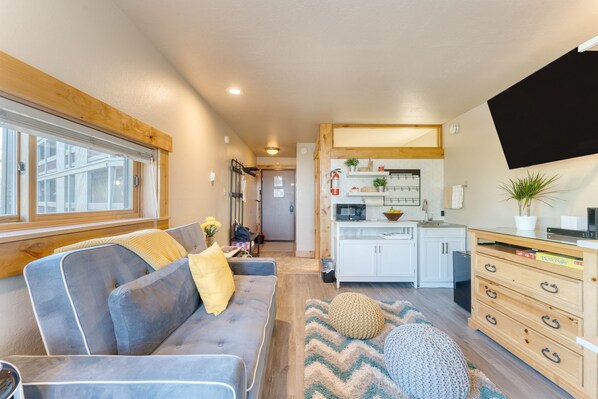 A modern living room with a sofa, a kitchen area with cabinets and a TV, and a hallway leading to another room. - Village Studio by Avantstay Minutes to Lifts (Truckee)