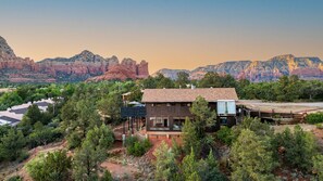 A modern house with a large deck, surrounded by dense greenery and red rock formations in the background. - Roca Roja by Avantstay Mountain Views in the Heart of Sedona (Sedona)