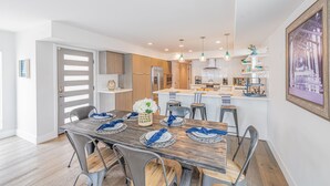 A modern kitchen with a central island, pendant lighting, and a dining area with a wooden table set for six. - Seaside Style by Avantstay Rooftop Walk to Beach Chic Design (Newport Beach)