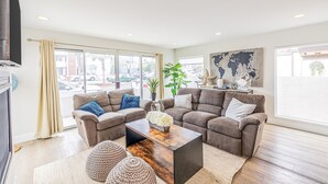 A spacious living room with a sectional sofa, a coffee table, and large windows offering a view of the street. - Seaside Style by Avantstay Rooftop Walk to Beach Chic Design (Newport Beach)
