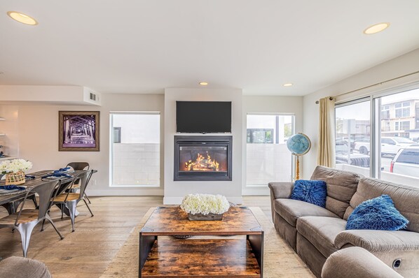 A modern living room with a fireplace, a dining area, and a large window offering a view of the city. - Seaside Style by Avantstay Rooftop Walk to Beach Chic Design (Newport Beach)