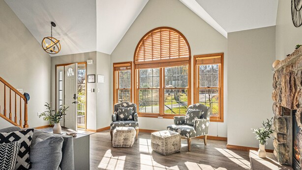 A spacious living room with a high ceiling, large windows, a fireplace, and a staircase.