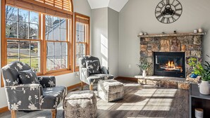 A living room with a fireplace, a clock, and a large window with blinds.