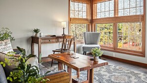 A cozy room with a wooden dining table, a comfortable chair, a desk with a lamp, and large windows with bamboo shades.