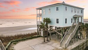 A light blue two-story house with a balcony, wooden deck, and a staircase leading to the upper floor, situated on stilts over a sandy beach with a view of the ocean. - Poseidon By Avantstay On The Beach Two Ocean Facing Balconies Hot Tub (Folly Beach)