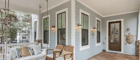 A covered porch with a white swing set, two rocking chairs, and a brick floor.