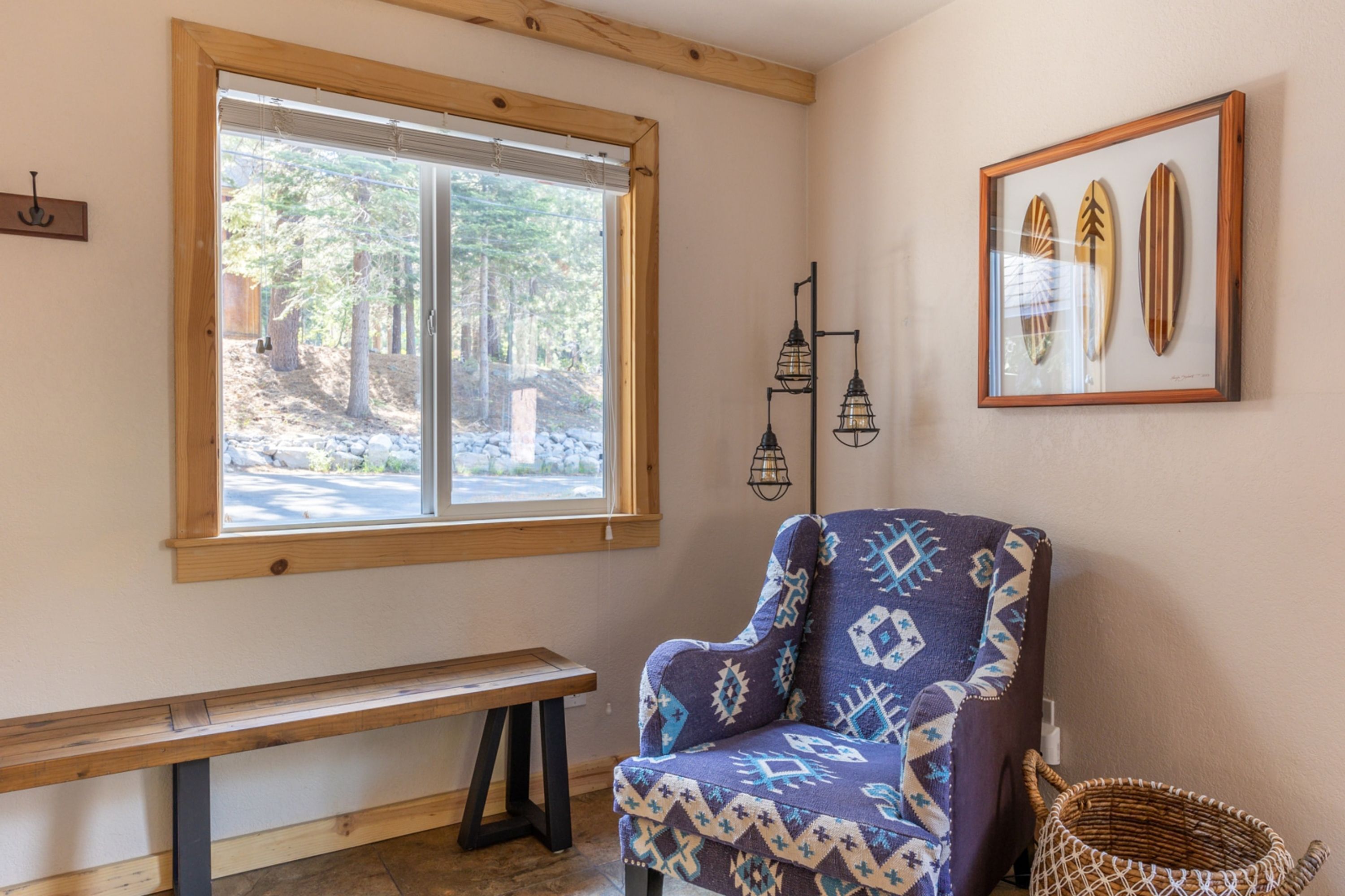 A room with a large window, a bench, a patterned armchair, a wall-mounted lamp, and a framed surfboard art piece.