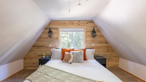 Un dormitorio con techo inclinado, una cama grande, dos mesitas de noche, un ventanal y paredes de madera.