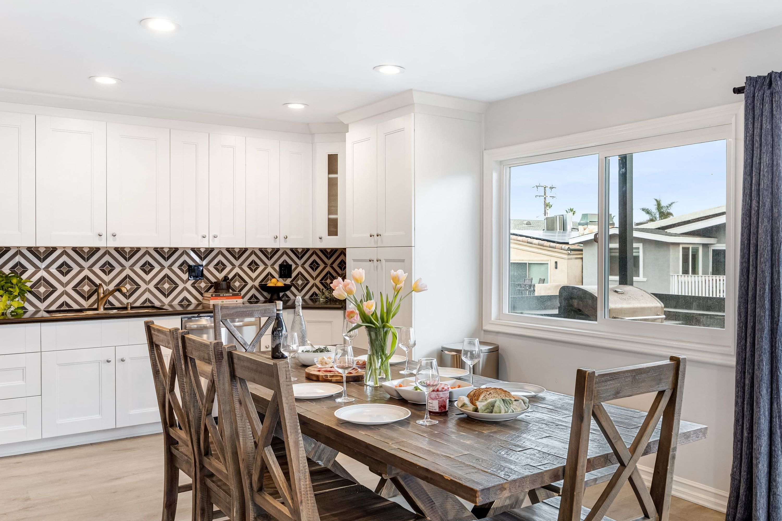 A modern kitchen with white cabinets, a wooden dining table, and a view of the neighborhood through a large window.