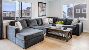 A modern living room with a grey sectional sofa, a wooden coffee table, and large windows offering views of neighboring houses. - Shelly by Avantstay Steps to the Beach Deck w Ocean Views 5bdr (Newport Beach)