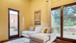 Ein Wohnzimmer mit einem beigen Ecksofa, einem gemusterten Teppich und einem großen Fenster mit Blick auf Grünflächen.
