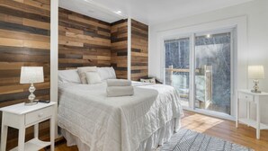 A modern bedroom with a large bed, wooden panel walls, a balcony with a view, and a white nightstand with a lamp. - Creekside Haven Gatlinburg Escape by a Bubbling Creek Near Top Attractions (Gatlinburg)