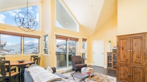 A spacious living room with a high ceiling, large windows offering a view of snow-capped mountains, a cozy seating area with a patterned rug, and a wooden cabinet. - Blue Mesa Penthouse by Avantstay Buyout of 3 Units Ski-in Ski-out Condo (Telluride)