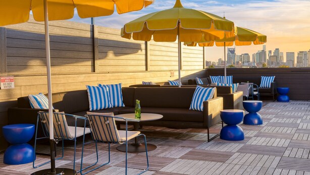 A rooftop lounge with outdoor seating, yellow umbrellas, and a city skyline view.