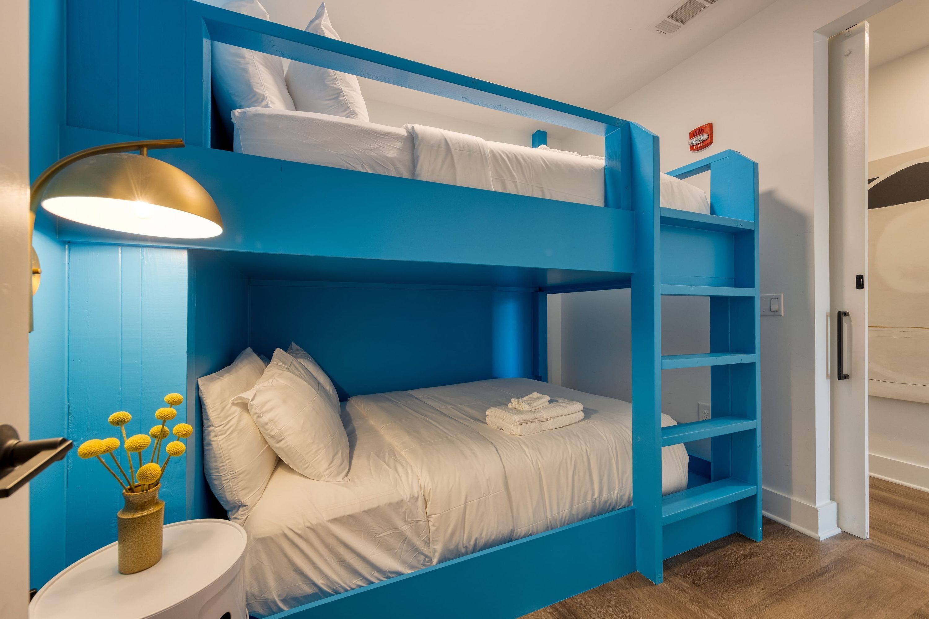 A bunk bed room with a blue color scheme, a bedside table with a lamp, and a vase with yellow flowers.