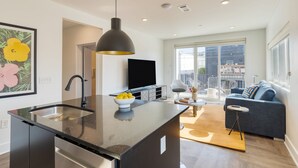 A modern kitchen with a black countertop and stainless steel appliances, a black pendant light, and a bowl of fruit on the counter.