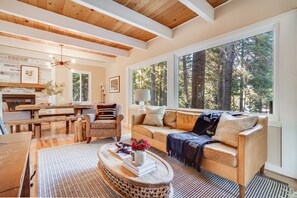 A cozy living room with a wooden ceiling, large windows with a forest view, a sofa, a coffee table with books and flowers, and a fireplace. - Homewood by Avantstay Amazing Family Cabin w Hot Tub Sauna (Homewood)