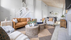 A modern living room with a staircase, a sofa, two round wicker ottomans, and a dining area. - Black Oak by Avantstay Modern Chic Santa Rosa Hideaway With Pool Table Fire Pit (Santa Rosa)