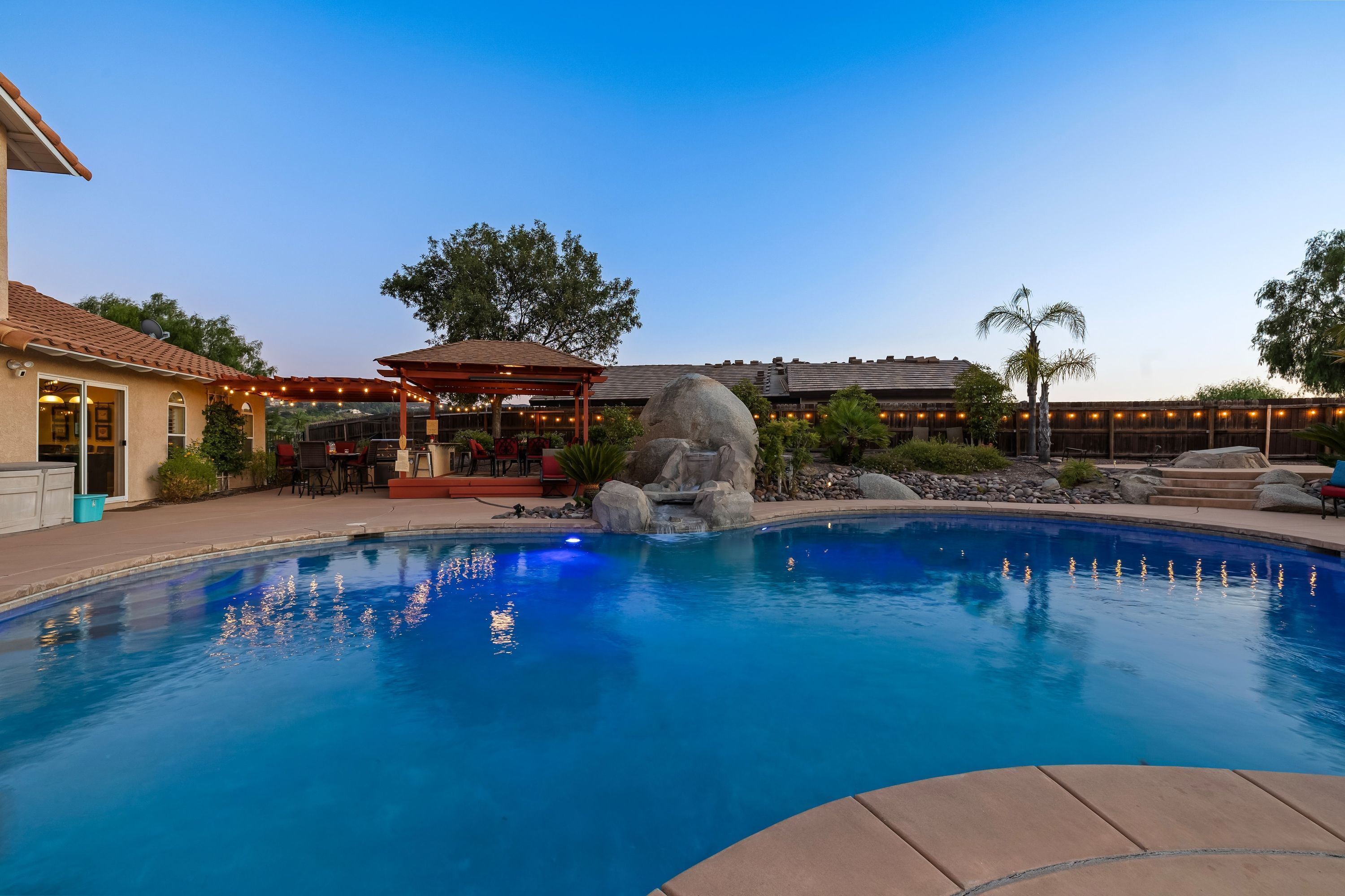 A swimming pool with a rock feature, a covered patio, and a building with outdoor lighting.