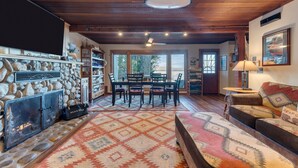 A cozy living room with a stone fireplace, a dining area with a table and chairs, and a large window offering a view of the outdoors. - Lakefront Haven by Avantstay Minutes to Ski Resorts Private Dock Boat (Homewood)