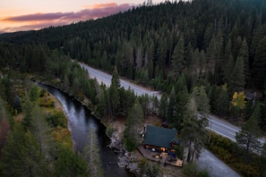 Eine Holzhütte an einem Fluss, nahe einer Straße, umgeben von dichtem Wald.