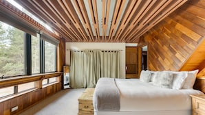 A bedroom with a large bed, wooden ceiling beams, a window with curtains, and a view of the outdoors.