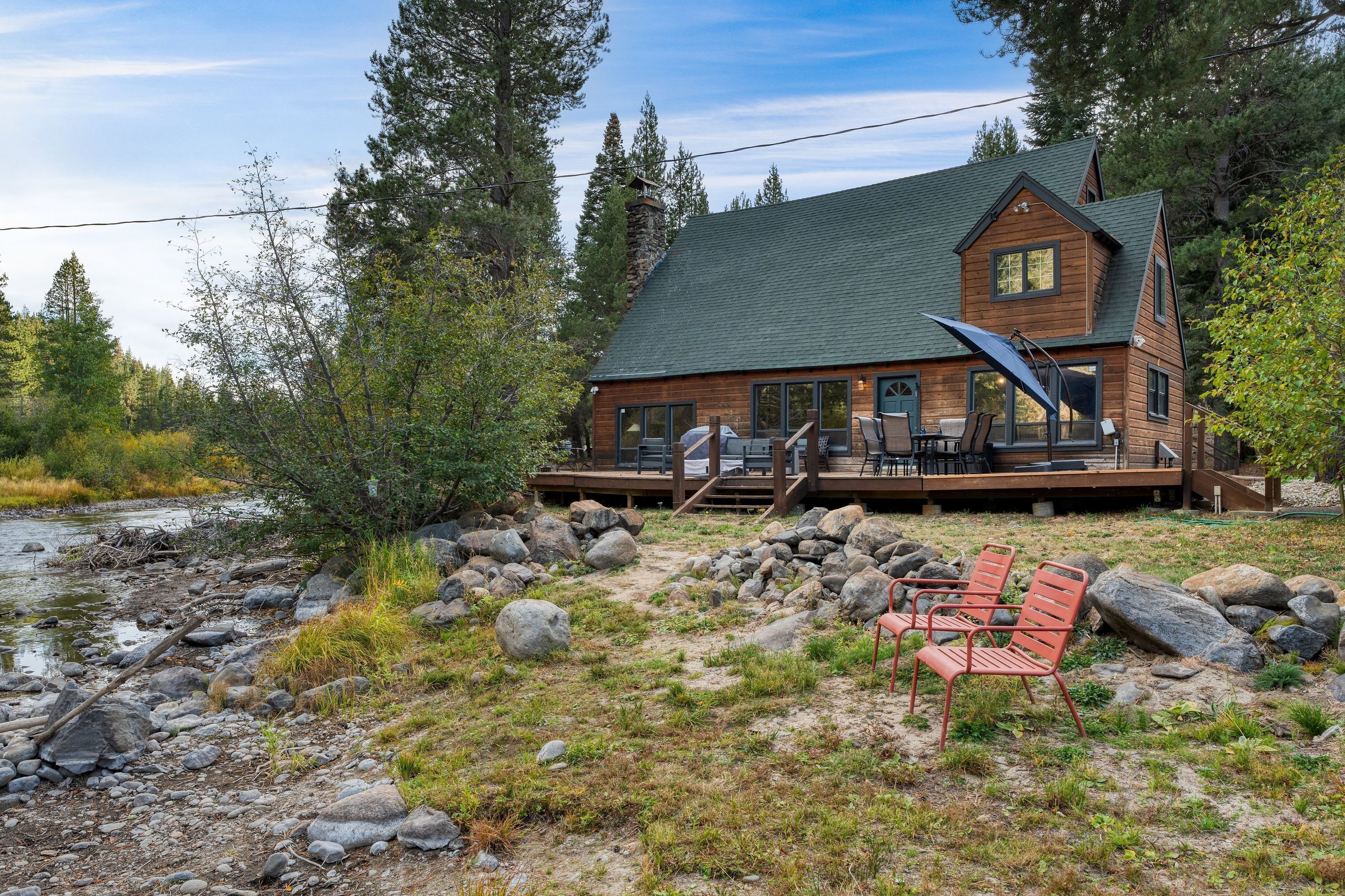 A cabin with a deck, surrounded by a natural landscape with a river and trees.